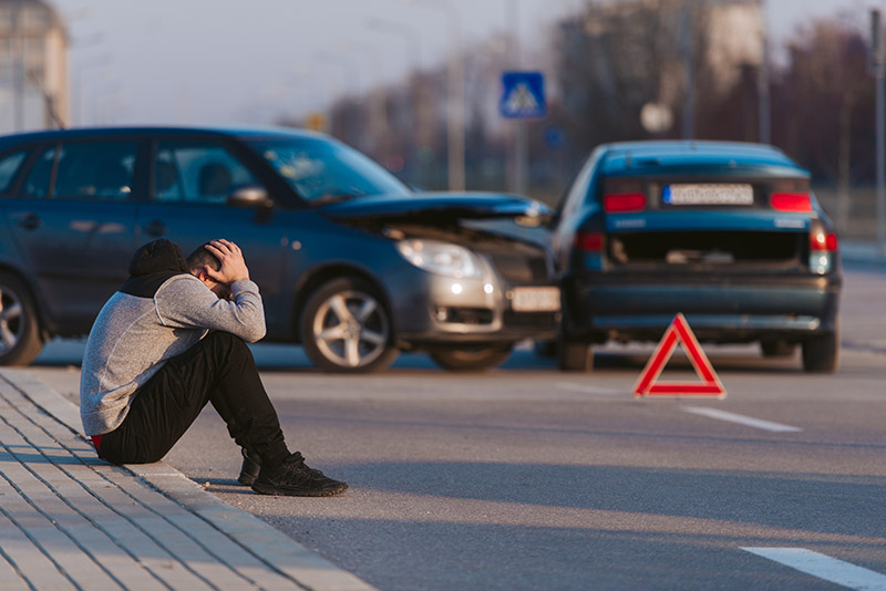 Mann sitzt nach einem Unfall an der Straße. Im Hintergrund zwei kollidierte Autos mit Warndreieck. 